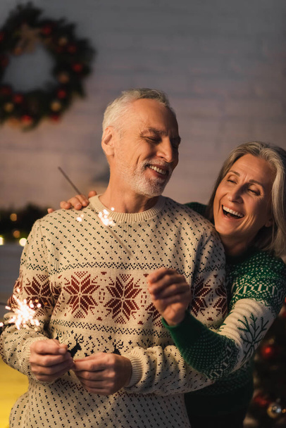 positive femme d'âge moyen étreignant mari barbu tout en tenant scintillant la veille de Noël - Photo, image