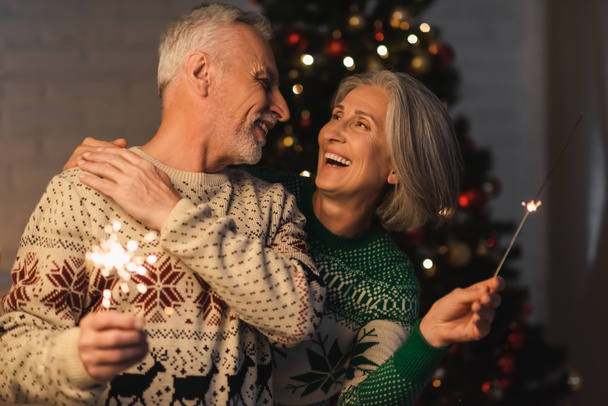 positive middle aged woman hugging bearded husband while holding bright sparkler on christmas eve - Photo, Image