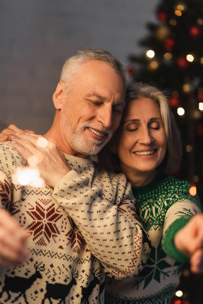 positive middle aged woman hugging bearded husband with closed eyes while holding sparkler on christmas eve - Valokuva, kuva