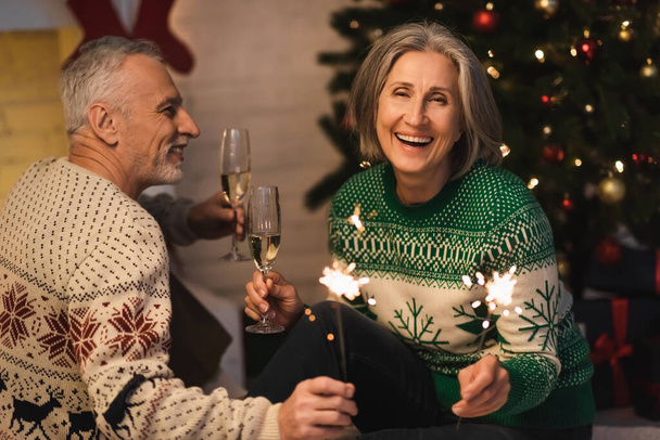 positive middle aged couple holding glasses of champagne and shiny sparklers on christmas eve  - Fotoğraf, Görsel