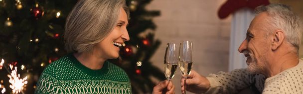 sonriente pareja de mediana edad tintineo vasos de champán cerca de brillante chispeante en la víspera de Navidad, bandera - Foto, Imagen