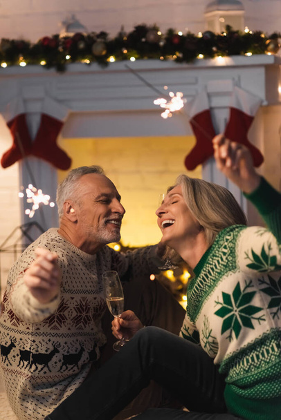 cheerful mature couple holding glass of champagne and shiny sparklers on christmas eve  - Foto, Bild