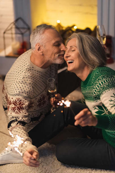 positive middle aged man kissing wife and holding shiny sparklers on christmas eve  - Photo, Image