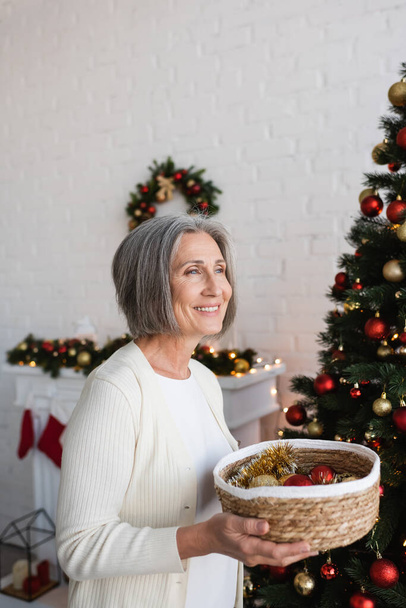 mujer feliz y madura sosteniendo cesta de mimbre cerca del árbol de Navidad decorado en casa - Foto, imagen