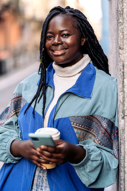 Beautiful african woman in the street using her phone and holding coffee cup. She is smiling and looking at camera. - Фото, изображение
