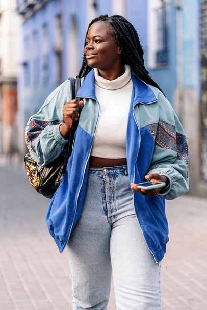 Pretty african american woman with cool braids walking in the city and using her mobile phone. - Foto, Imagen
