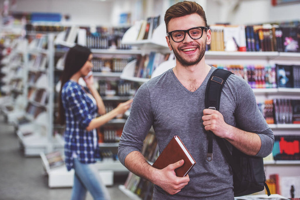 Estudante bonito em óculos está segurando livro, olhando para a câmera e sorrindo enquanto está de pé na livraria, no fundo menina está escolhendo livro
 - Foto, Imagem