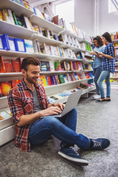 Estudiante guapo está utilizando un ordenador portátil y sonriendo mientras está sentado en la librería, en el fondo las niñas están eligiendo libros
 - Foto, Imagen