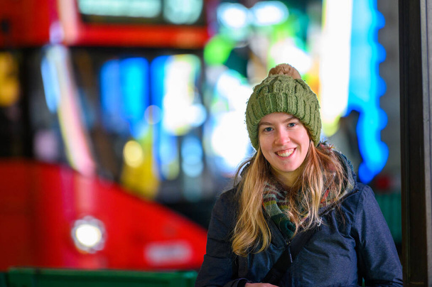 Kırmızı Londra çift katlı otobüsü olan, oyuncak şapkalı gülümseyen sarışın kadın ve arka planda odak noktasından çıkan neon ışıkları. - Fotoğraf, Görsel