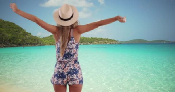 Achteraanzicht van stijlvolle vrouw met uitgestrekte armen die wind voelen op tropisch strand. Aantrekkelijke jonge Kaukasische dame in zomerkleding genietend van de strandsfeer. 4k - Video
