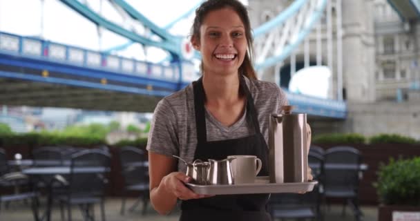 Pěkná servírka nesoucí podnos s kávou poblíž Tower Bridge. Přátelská mladá žena v Anglii, v zástěře a s podnosem na servírování kávy. 4k - Záběry, video