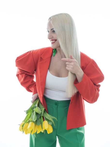 Eine modische Momentaufnahme. Eine schöne Blondine posiert in heller Kleidung vor weißem Hintergrund mit gelben Tulpen in den Händen. Schönheit, Modekonzept. - Foto, Bild