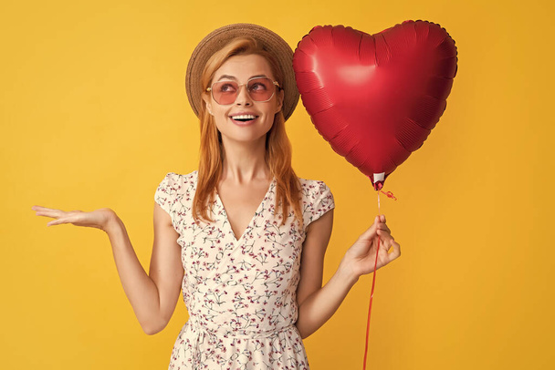 щаслива молода жінка в солом'яному капелюсі і сонцезахисних окулярах тримає любовну повітряну кулю на жовтому тлі. представлення продукту
. - Фото, зображення