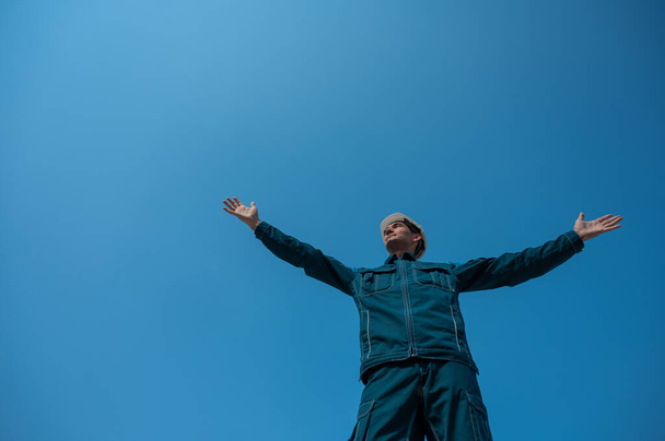 A caucasian man in work clothes and a construction helmet stands against the background of a blue sky with his arms outstretched to the sides - Photo, image