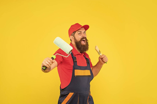 angry bearded man in work clothes hold paint roller and brush on yellow background. - Foto, afbeelding