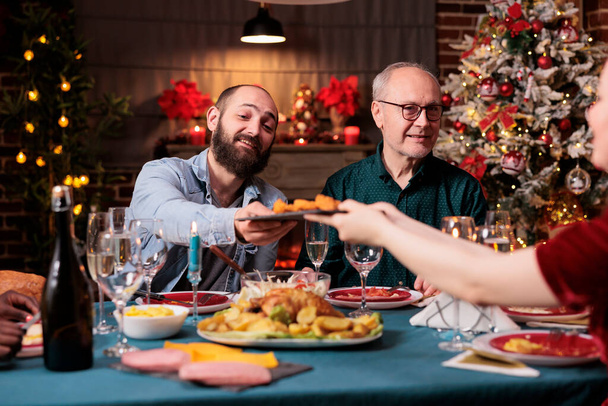 一緒にクリスマスディナーをしたり、お祝いのテーブルで伝統的な食べ物を食べたり、女性プレートを皿で渡したりする家族。冬休みに両親と一緒に集まり、クリスマスを祝う - 写真・画像