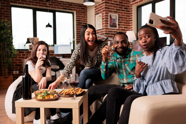 Multi ethnic young adults group posing for mobile selfie at apartment living room, smiling, laughing, tasting snacks drinking wine. African american woman photographing friends. - Photo, Image