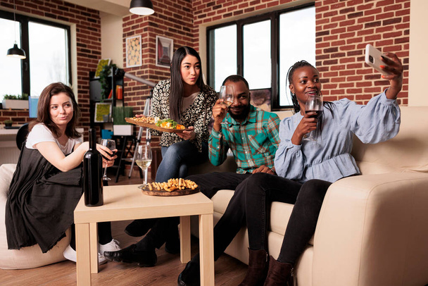 Africano americano, asiático, caucásico adultos jóvenes tostando vasos de vino, sosteniendo snack board, sonriendo para la fotografía del teléfono inteligente durante la fiesta de la casa en la sala de estar del apartamento. - Foto, imagen