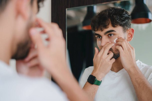 Egy fehér pólós férfi portréja, aki foltokat visel a szeme alatt, miközben az arcbőrét ápolja, tükörbe néz. Szépségápolás. Bőrgyógyászat, kozmetika. Szemészeti ellátás - Fotó, kép
