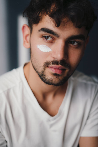 Man portrait of a handsome young man applying moisturising cream to his face, looking away. Beauty portrait. Man portrait. Beauty routine. Skin care routine. - Foto, afbeelding