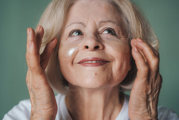 Közelkép egy nyugdíjas nőről, aki kozmetikai beavatkozásokat végez, szemkötőkkel a szeme alatt, zöld fal felett. Száraz bőrápolás. Szépségterápiás koncepció - Fotó, kép