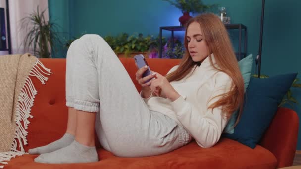 Muotokuva valkoihoinen aikuinen tyttö käyttää älypuhelimen kirjoittamalla selailu, menettää tulossa yllättynyt äkillinen arpajaiset tuloksia, huonoja uutisia, onni menetys, epäonnistua. Nuori nainen makaa oranssi sohvalla kotona olohuone - Materiaali, video