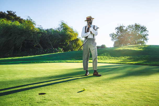 View of a fashionable handsome bearded black senior with a sigar, in straw hat and tailored outfit standing on a green golf field with a club in his hand; a yellow golf ball and a hole in front of him - Фото, изображение