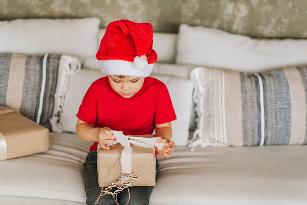 Happy laughing Caucasian boy in red Santa Claus hat with his presents on Christmas morning. Child excited and smiling. Happy family holidays at home. Opening gifts in boxes. Copy space. High quality - Photo, Image