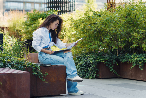 Concentrated young female entrepreneur in casual clothes and glasses examining documents while sitting near office building in resting outdoor area. - Photo, image