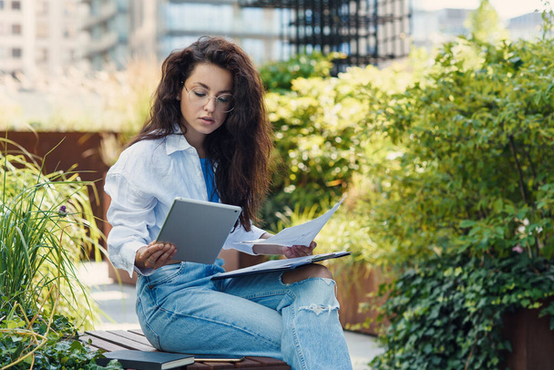Concentrated young female entrepreneur in casual clothes and glasses examining documents while sitting near office building in resting outdoor area. - Foto, immagini