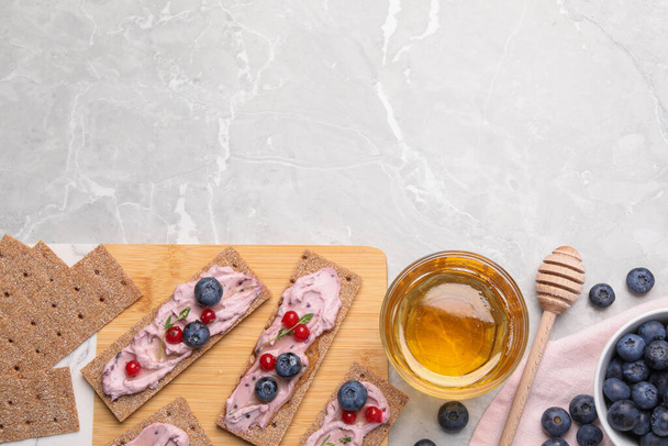 Tasty cracker sandwiches with cream cheese, blueberries, red currants, thyme and honey on light grey marble table, flat lay. Space for text - Photo, image