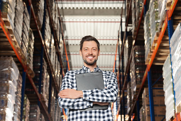 Happy manager holding clipboard in warehouse with lots of products, low angle view - Zdjęcie, obraz