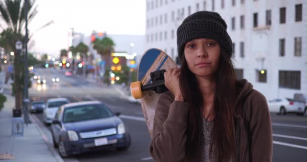 Millennial κορίτσι κρατώντας skateboard πάνω από τους ώμους της κοιτάζοντας κάμερα στο δρόμο, όπως ο ήλιος δύει. Μέτριο πορτραίτο μιας χίπστερ γυναίκας με στάση στο κέντρο το σούρουπο. 4ια - Πλάνα, βίντεο