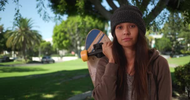Millennial dívka drží skateboard přes rameno dívá se na kameru v parku během dne. Střední portrét hipsterky s postojem stojící pod stromem. 4k - Záběry, video