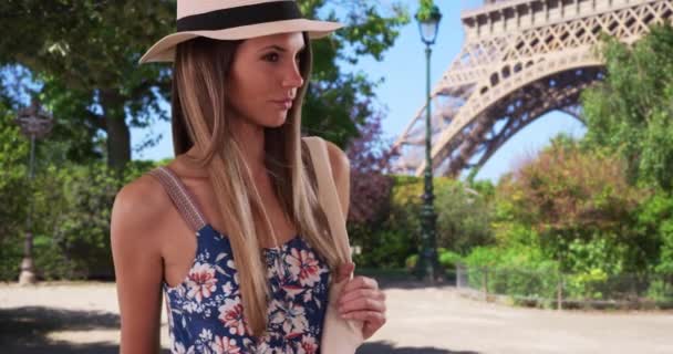 Pretty lady in floral romper wearing fedora and holding bag near Eiffel Tower. Stylish young woman in her 20s in summer dress standing casually in Paris, France. 4k - Footage, Video