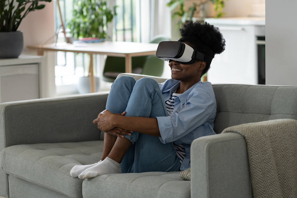 Young joyful African woman wearing modern VR headset sitting on sofa in living room, watching immersive 3D video, overjoyed black girl using virtual reality glasses at home. Future technologies - Photo, Image