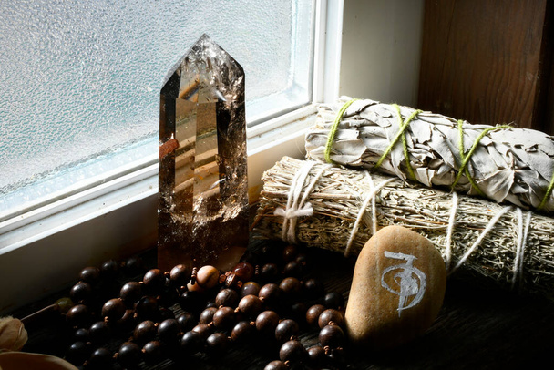 Λευκό φασκόμηλο μουτζούρες με ένα καπνιστό πύργο κρύσταλλο χαλαζία και χάντρες mala κοντά σε ένα φωτεινό παράθυρο.  - Φωτογραφία, εικόνα