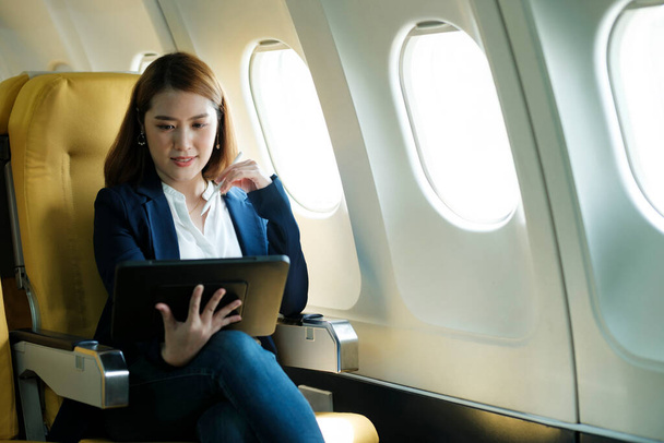 Joven mujer de negocios asiática profesional en ropa formal trabajando con tableta con lápiz inteligente mientras está sentado en cabina de avión cerca de la ventana que viaja a otro lugar. Concepto de viajes y negocios. - Foto, imagen