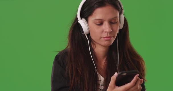 Jonge vrouwelijke student luisteren naar muziek met smartphone en koptelefoon op groen scherm. Schattig wit meisje dat sms 'jes leest terwijl ze naar muziek luistert op een smartphone op het groene scherm. 4k - Video