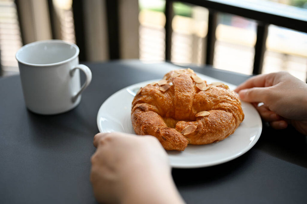 A female hands holding a delicious fresh French croissant with almond slice on a white plate serve with hot coffee or cocoa. - Фото, изображение