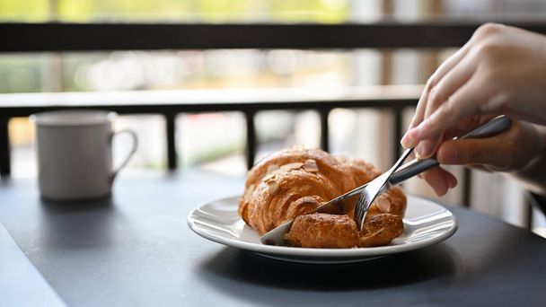 Una hembra comiendo un delicioso croissant francés fresco con rebanada de almendras en un plato blanco y café para su desayuno en la cafetería. imagen recortada - Foto, Imagen