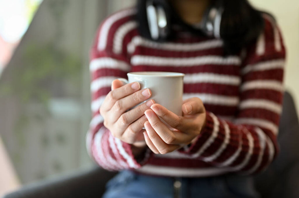 Mujer asiática joven con ropa casual sosteniendo una taza de café o té, relajándose en la cómoda cafetería. imagen recortada y de primer plano - Foto, imagen