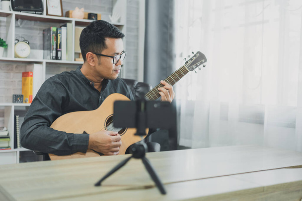Asian music teacher man broadcasting live guitar lessons to students online using mobile. Asian male musician playing guitar singing live video conferencing at home. Live concert music from anywhere. - Zdjęcie, obraz