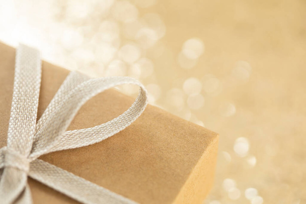 Close up fragment of the brown paper gift box with linen bow tie on blurred smimmering golden background. Selective focus. - 写真・画像