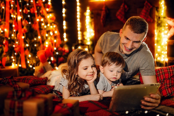 Father and his little daughter and son using a tablet pc at home by a fireplace in warm and cozy living room on Christmas eve. Winter evening at home with family and kids. - Foto, afbeelding