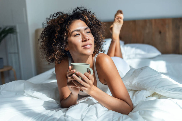 Shot of beautiful woman drinking a cup of coffee while staying on the bed.  - 写真・画像
