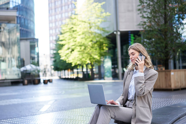 Бізнес-леді робить дзвінки і працює на відкритому повітрі, сидячи в центрі міста в костюмі, тримаючи ноутбук і мобільний телефон
. - Фото, зображення