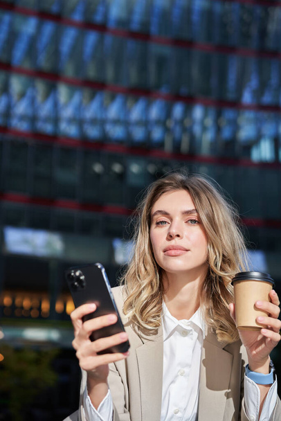 Stijlvolle jonge corporate vrouw staat in de straat, drinkt koffie en houdt mobiele telefoon. - Foto, afbeelding