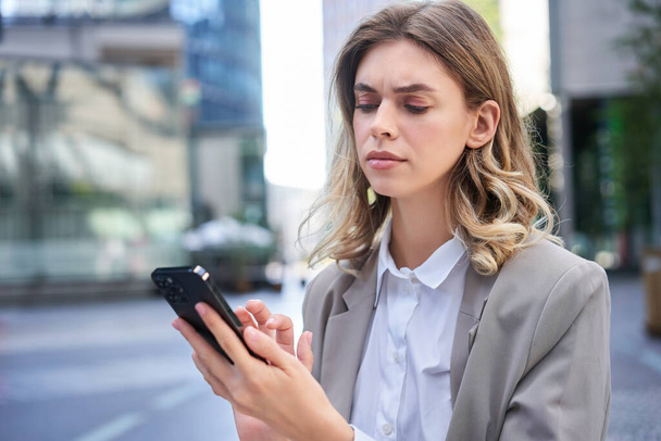 Young businesswoman looking complicated, frowning as reading message on mobile phone. Serious corporate woman checking her smartphone. - Photo, Image
