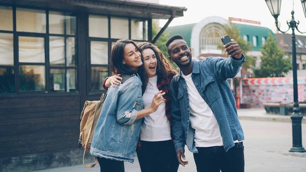 Happy African American guy is taking selfie with friends beautiful Caucasian girls standing in street posing and holding modern smartphone during journey in foreign country. - Fotografie, Obrázek